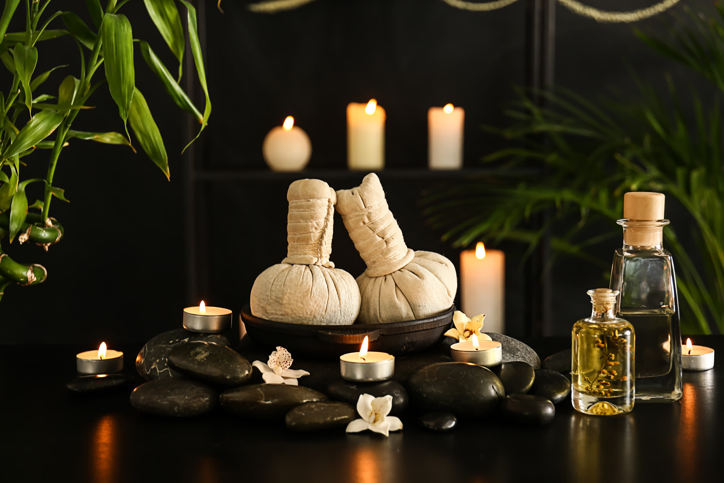 Spa still life with lit candles, hot stones, herbal compresses and massage oil on a dark background