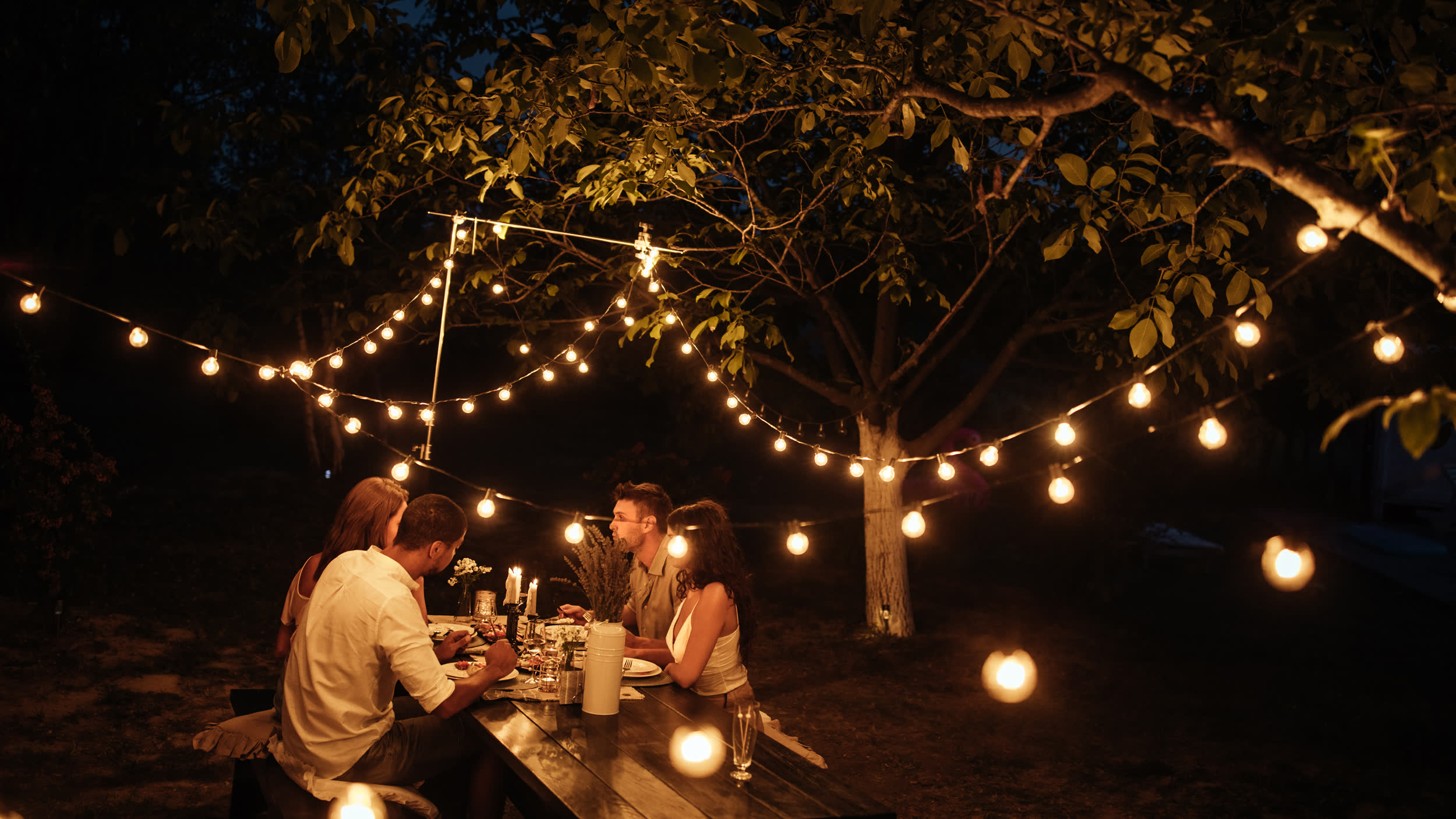 Guests dining under string lights beneath a tree on a summer evening